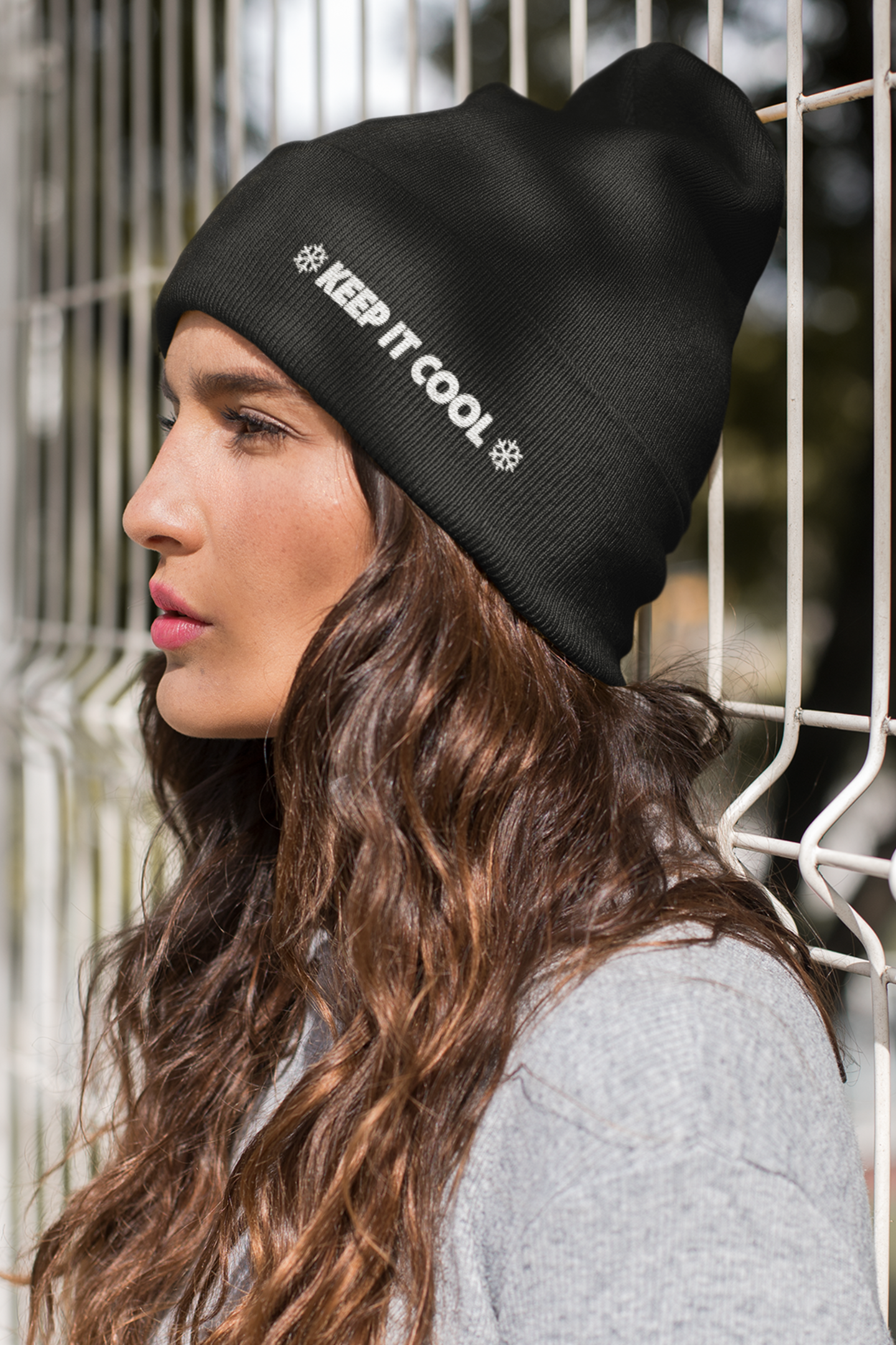 Woman wearing a black beanie with 'KEEP IT COOL' text, standing in front of a wire fence.