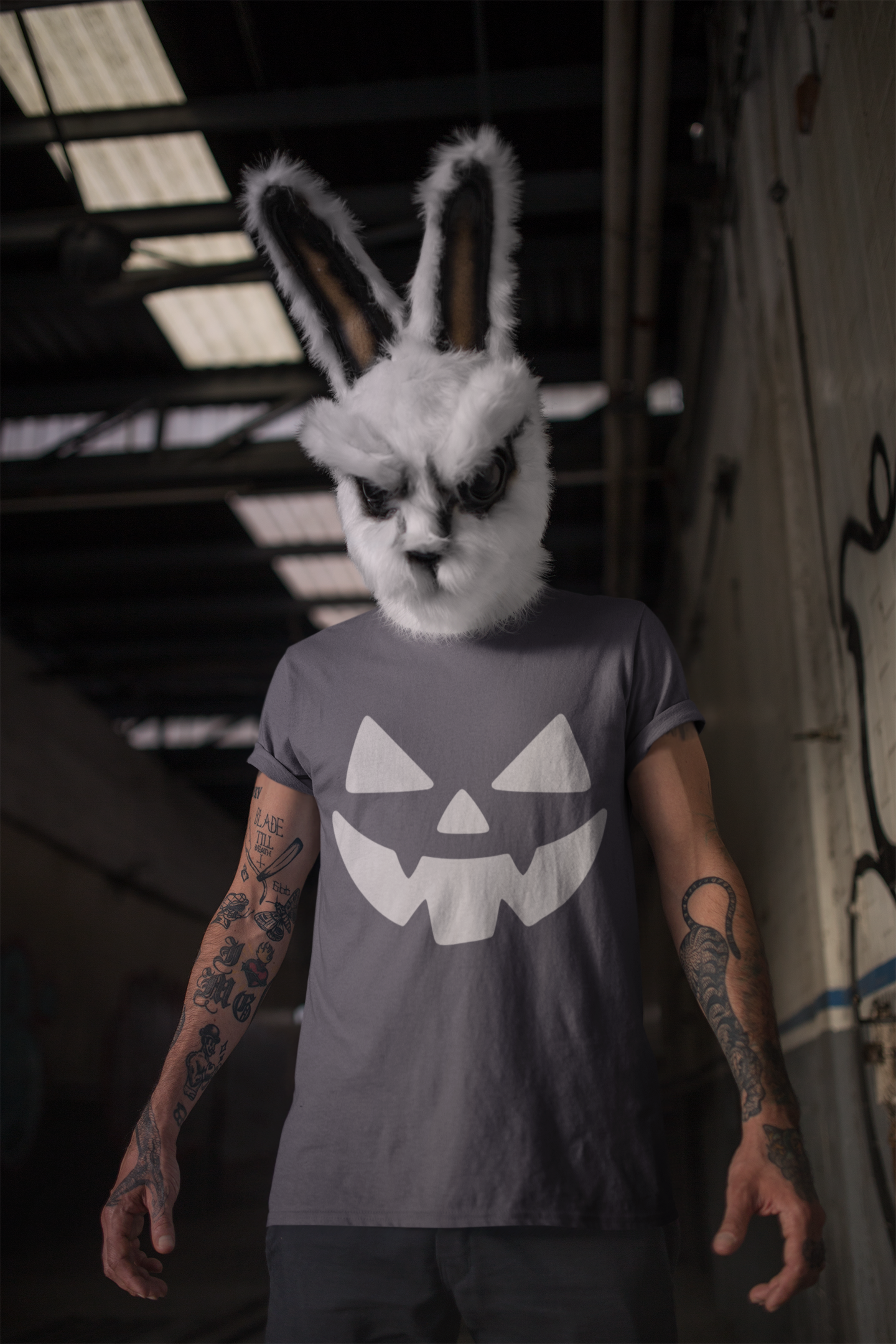 Person wearing a rabbit mask and a t-shirt with a jack-o'-lantern design in an industrial setting.