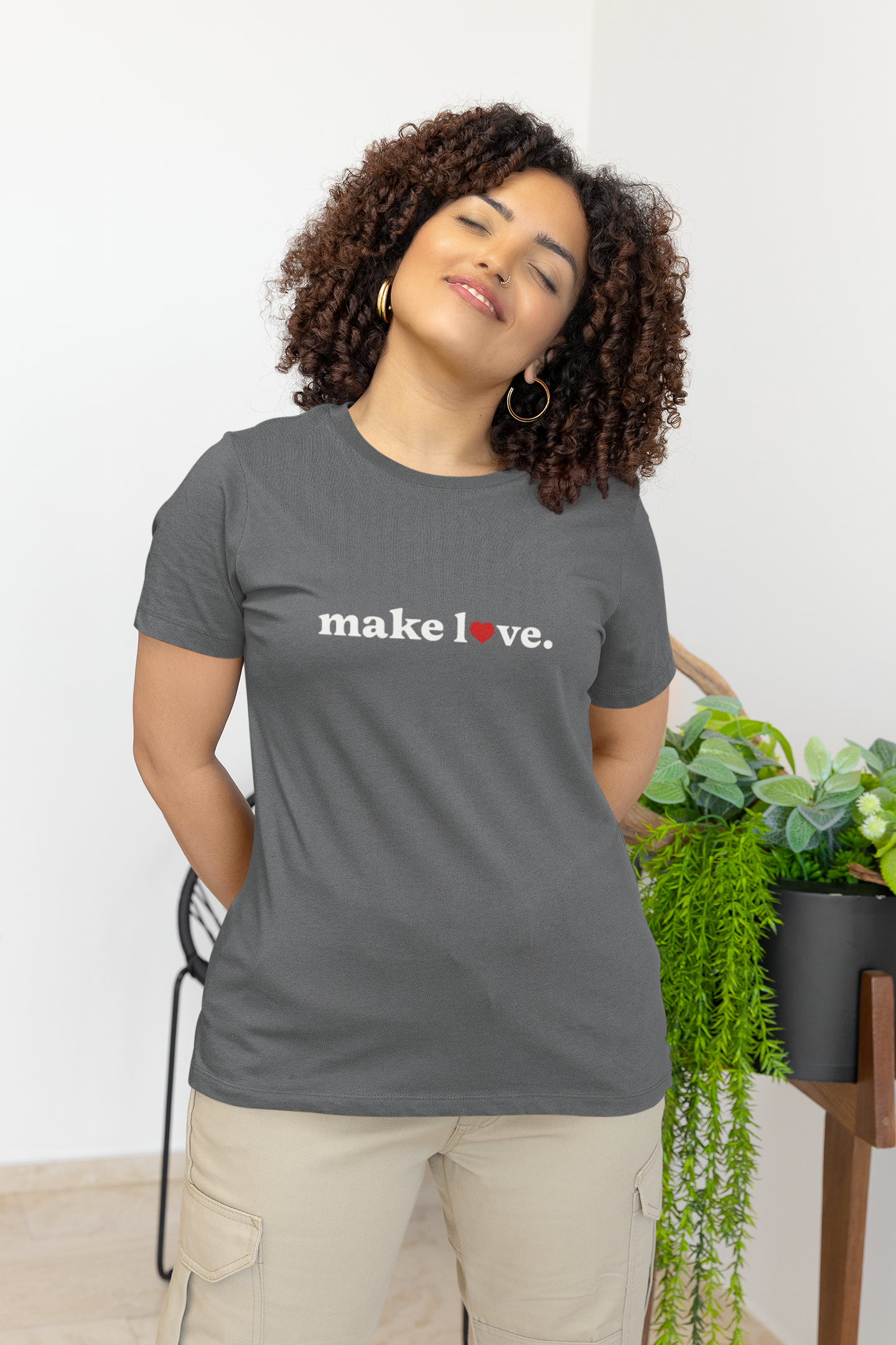 Woman wearing a gray t-shirt with 'make love' text, standing indoors with plants in the background.