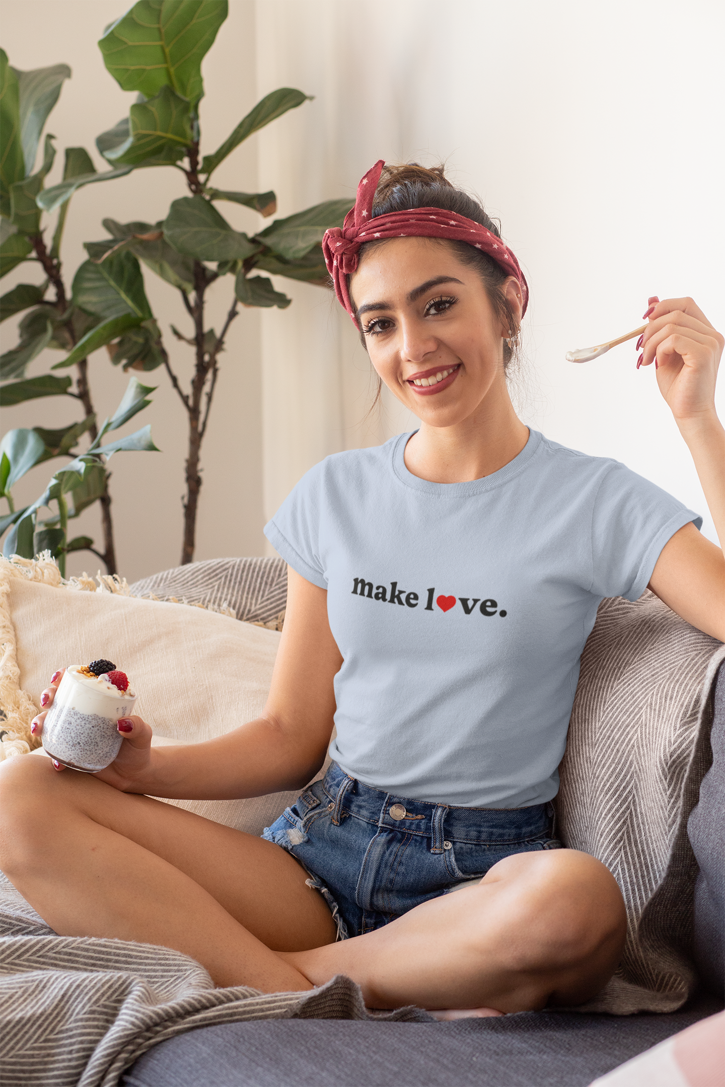 Woman sitting on a couch wearing a t-shirt with 'make love' text, holding a mug and a spoon.