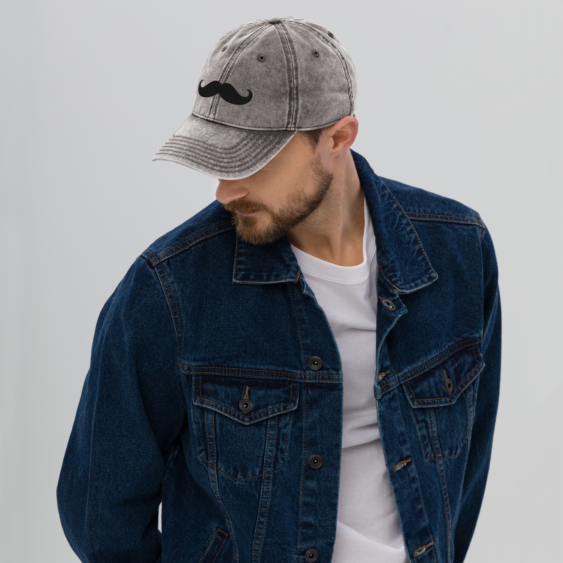Man wearing a denim jacket and cap with a mustache design on a plain background