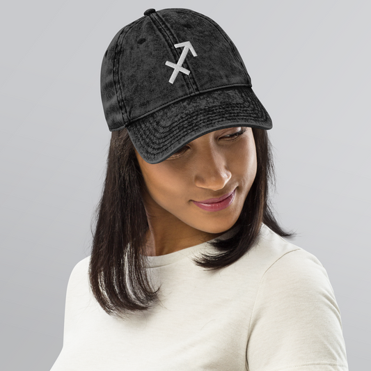 Person wearing a black baseball cap with a Sagittarius symbol on a gray background
