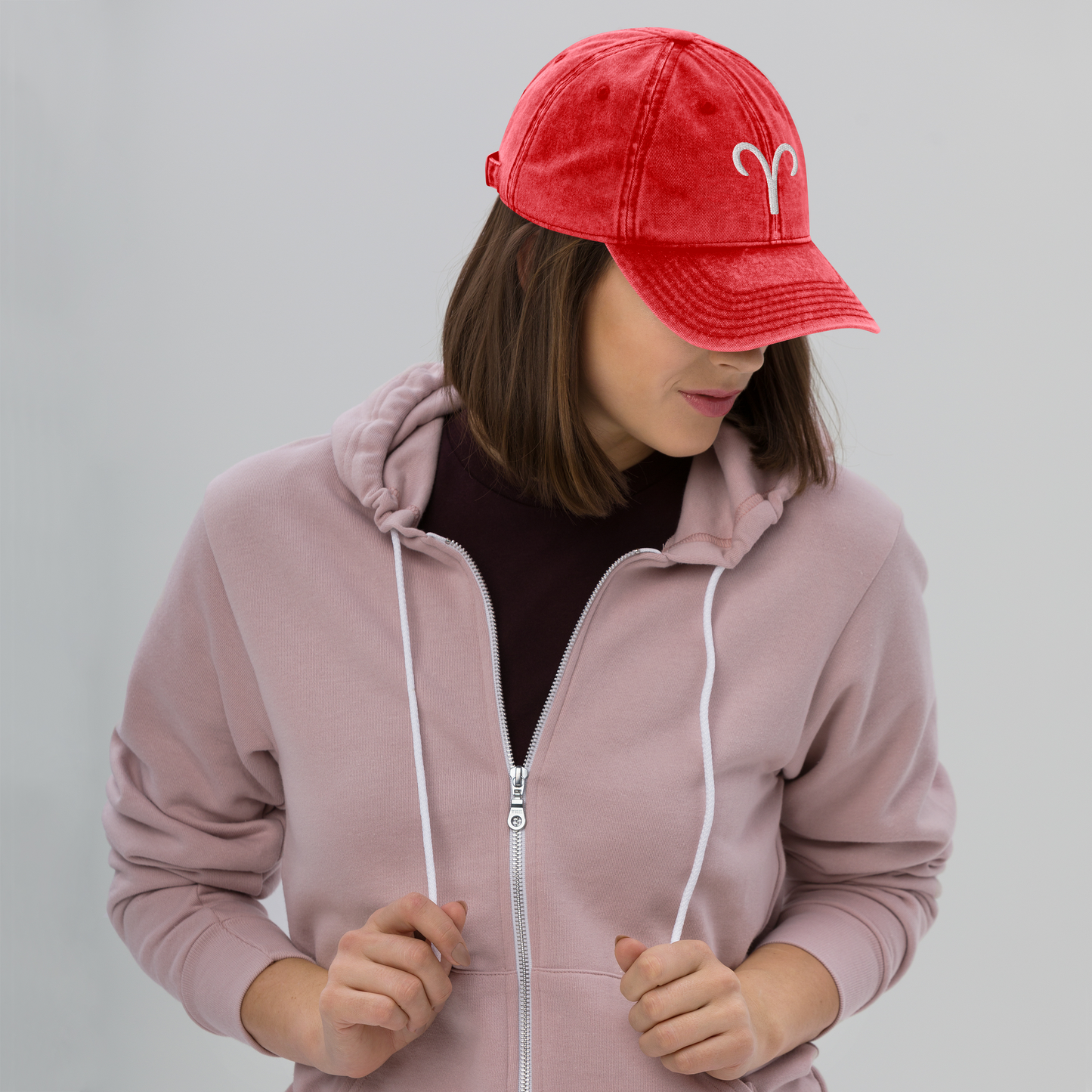 Person wearing a pink hoodie and red cap with a white aries logo on a gray background