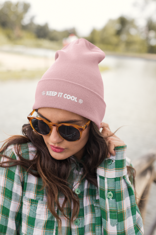 Woman wearing a pink beanie with 'Keep It Cool' text, sunglasses, and a green plaid shirt by a lake.