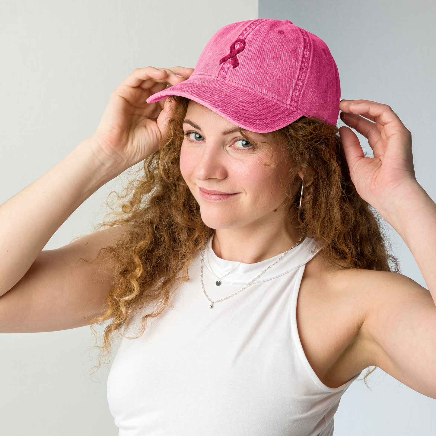 Woman wearing a pink cap with a ribbon symbol, standing against a plain background