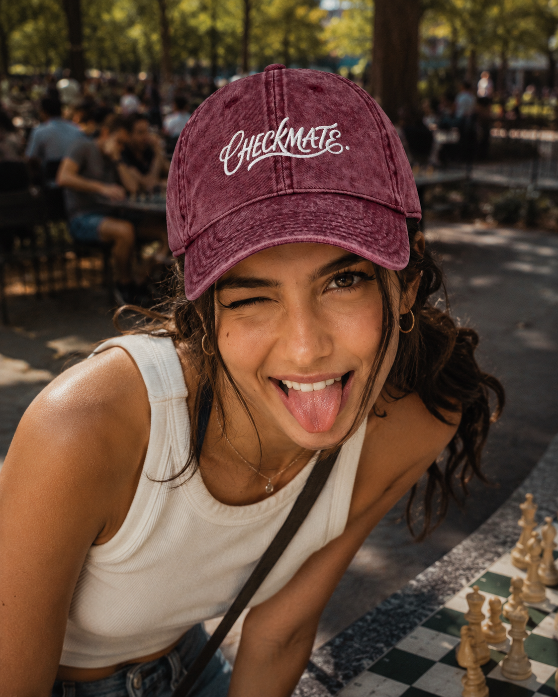 Woman wearing a maroon 'Checkmate' cap in an outdoor setting with people and trees.