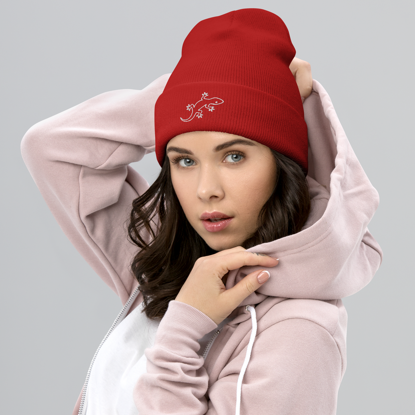 Woman wearing a red beanie with a white gecko design and a pink hoodie on a gray background