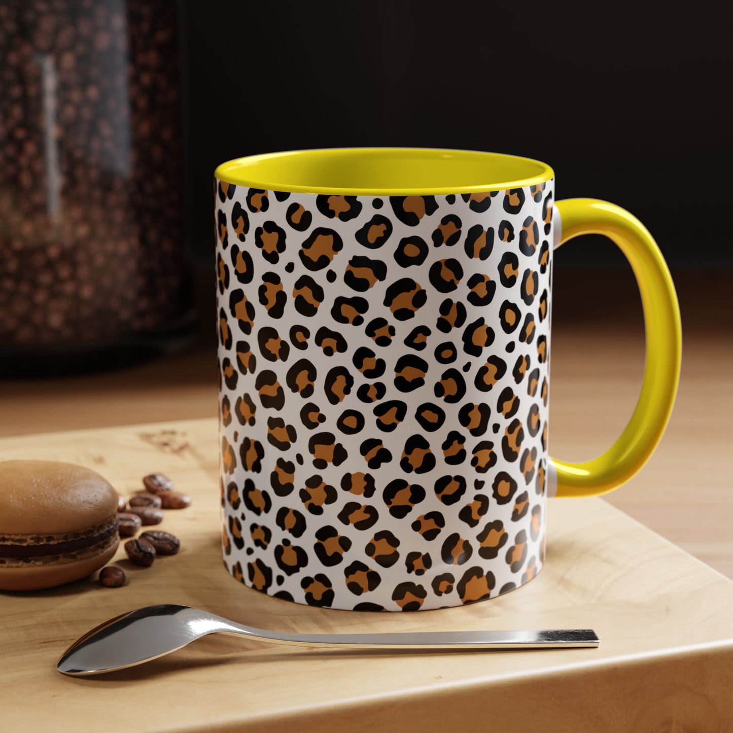 Mug with leopard print pattern and yellow handle on a wooden surface with coffee beans and a spoon.