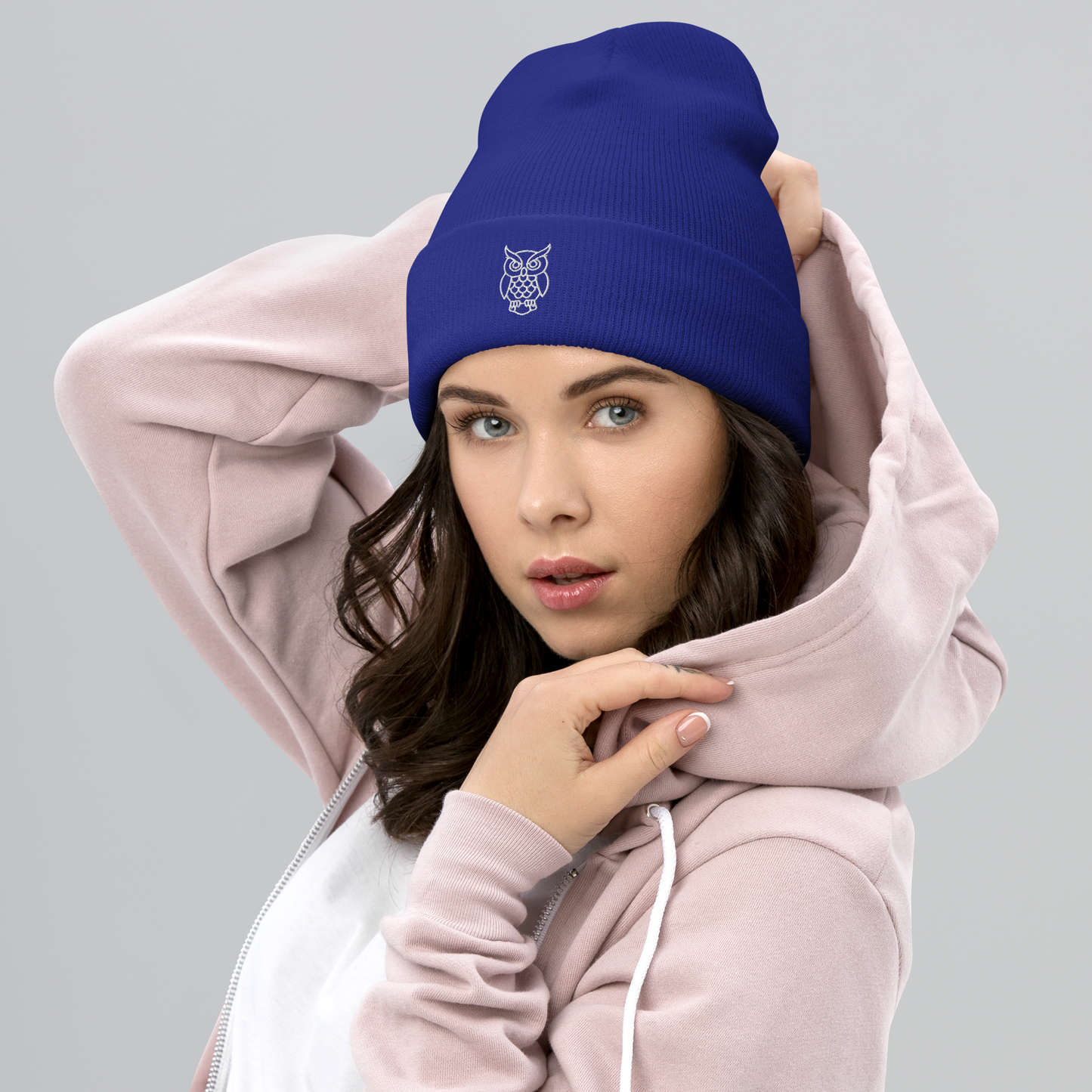 Woman wearing a blue beanie with an owl logo and a pink hoodie on a gray background