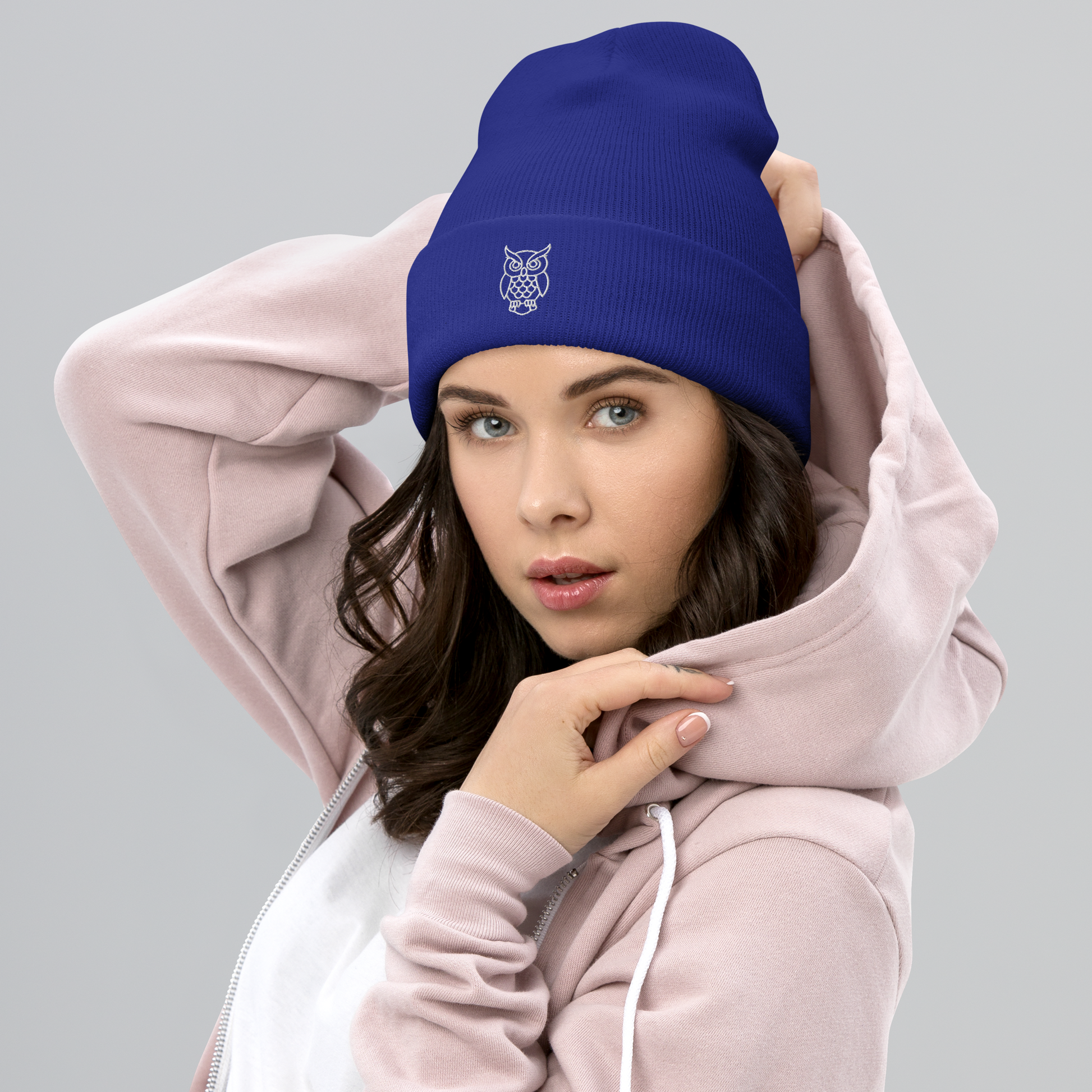 Woman wearing a blue beanie with an owl logo and a pink hoodie on a gray background