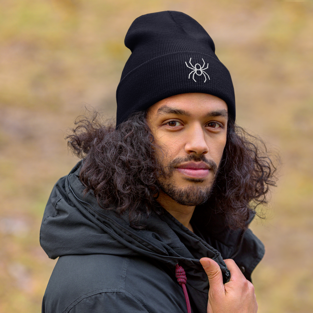 Man wearing a black beanie with a spider logo and a dark jacket outdoors.