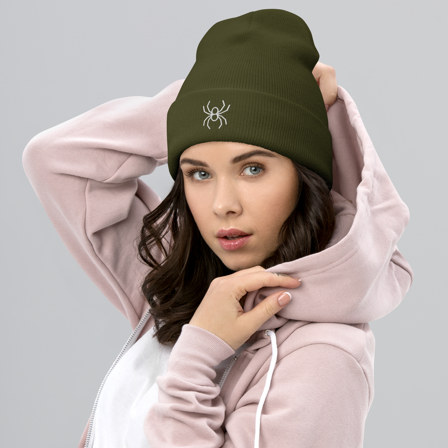 Woman wearing a green beanie with a spider logo and a pink hoodie on a gray background