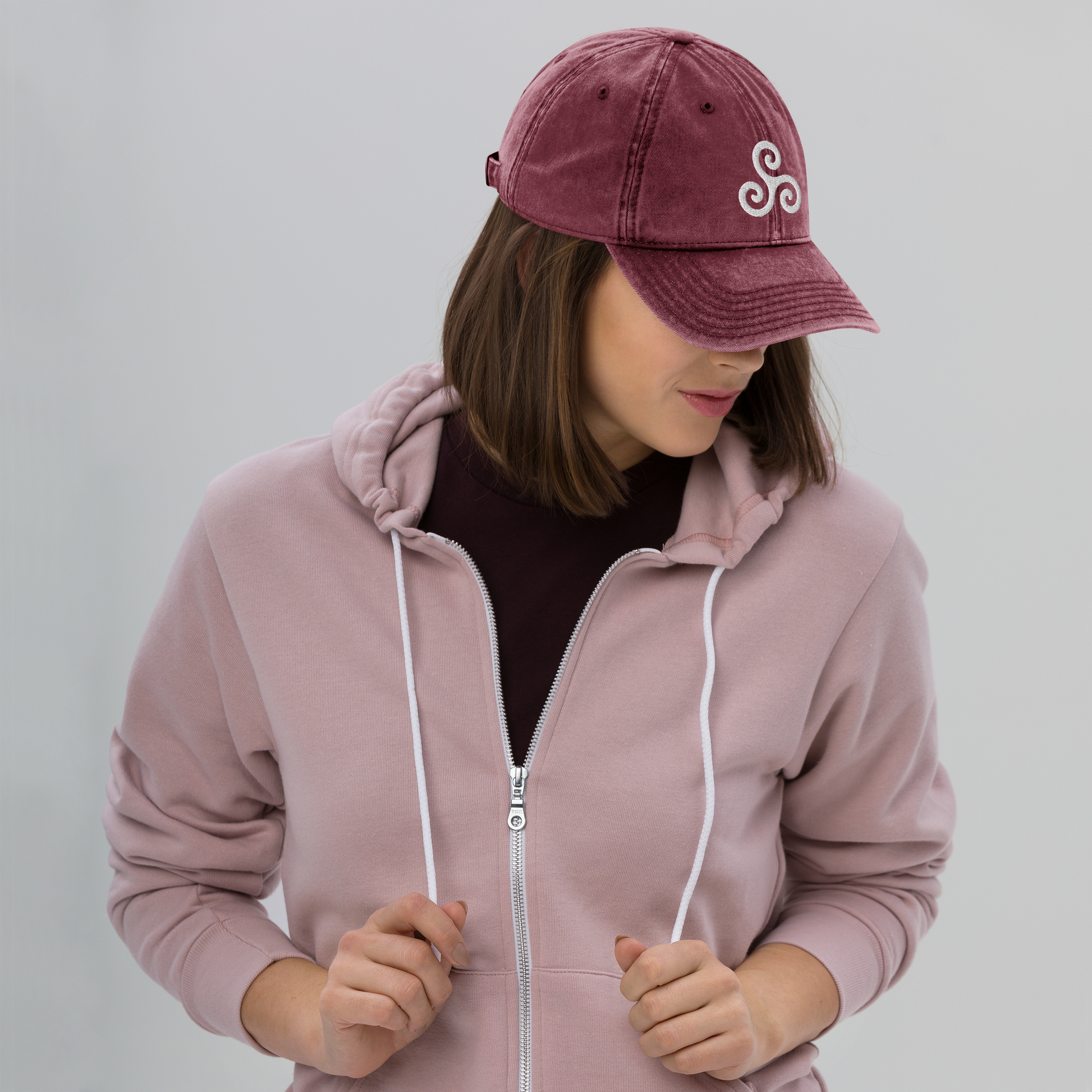 Person wearing a pink hoodie and maroon cap with a logo on a plain background